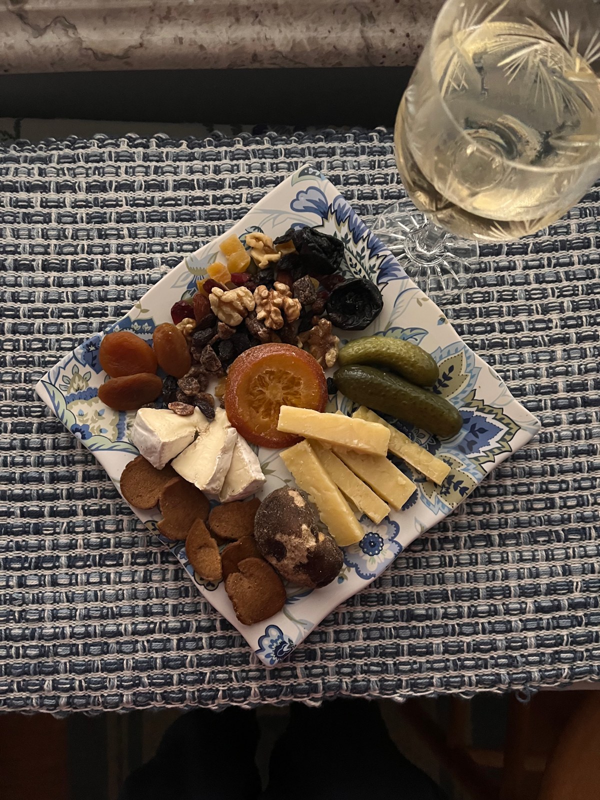 A blue-and-white plate with cheese, nuts, dried fruit, and pickles beside an etched stemmed glass on a woven placemat.