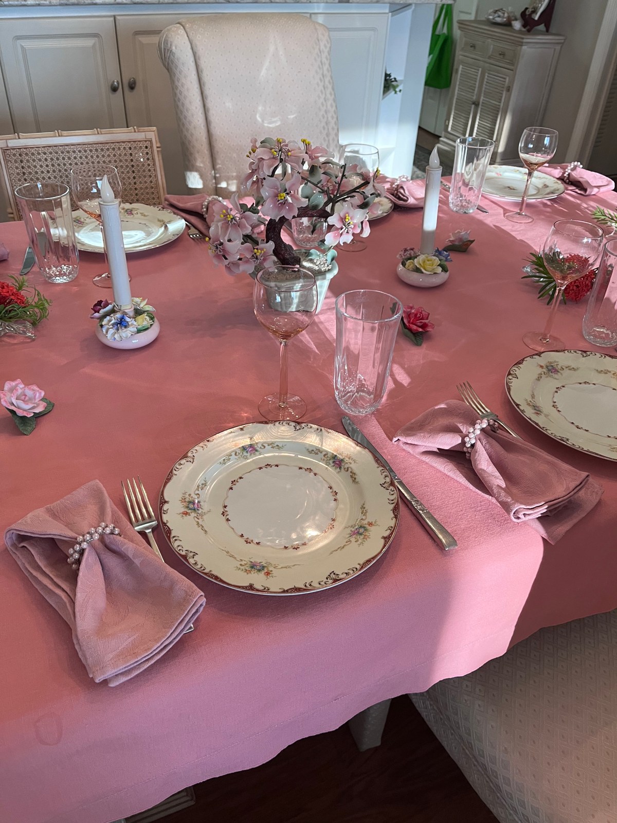 A pink dining table with floral china, pearl napkin rings, stemmed glasses, candles, and a flowering centerpiece.